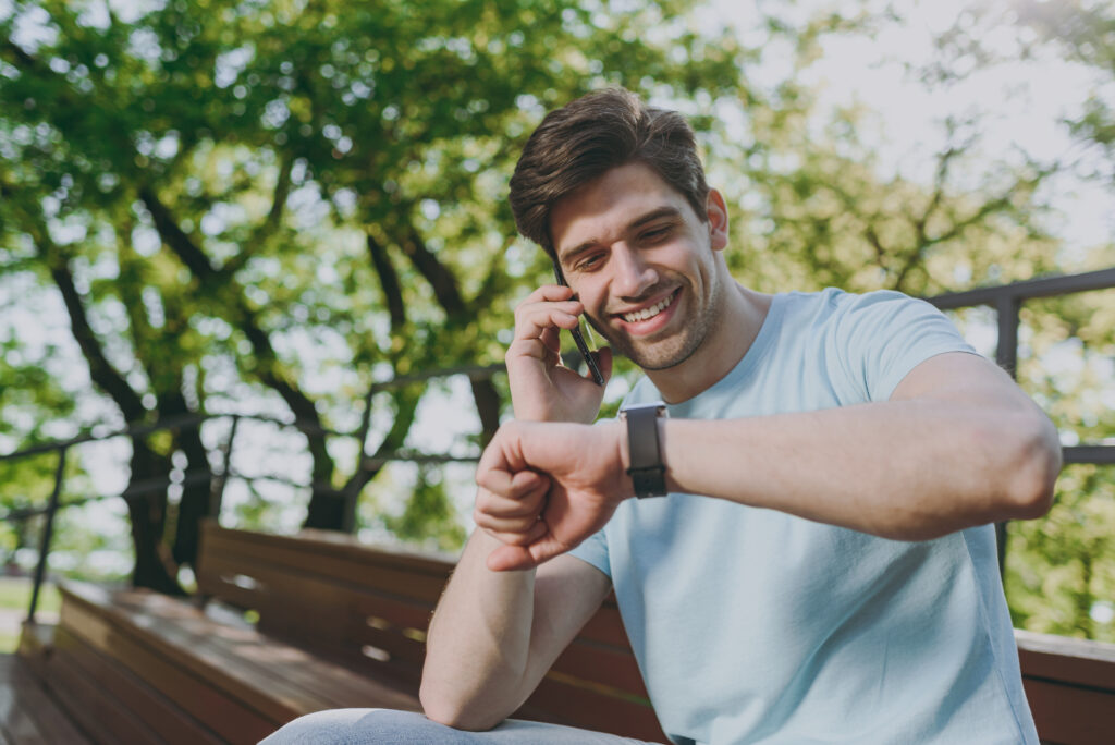 Smiling young happy man wear blue t-shirt sit on bench talk speak on mobile cell phone check time on smart watch rest relax in sunshine spring green city park outdoor on nature Urban leisure concept